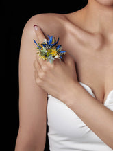 A person wearing a white top poses with the CAMELLIA Cancer Flower Ring, which features colorful dried flowers, on their right hand against their left shoulder.